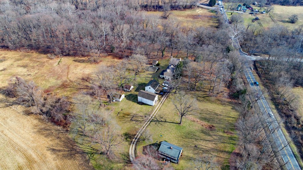 A view of the entire Cooch Homestead and surrounding landscape. A road runs along the right. The house and outbuildings are in the center, and farm fields to the left. The Christina River runs from the left to right above the buildings. The houses are bordered by trees and agricultural fields.