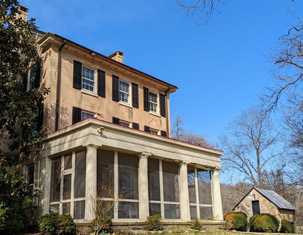 Front view of Cooch family house, beige two story building with large screened in porch supported by columns. Historic stone smokehouse in the background.