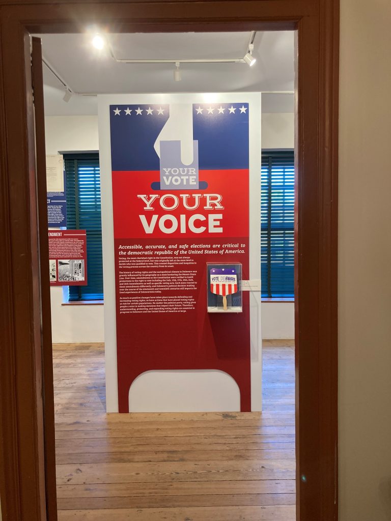 Entrance to the Your Vote, Your Choice exhibit; a large pillar with red white and blue coloring, featuring informational text about the exhibit and a promotional fan used for political campaigns. 