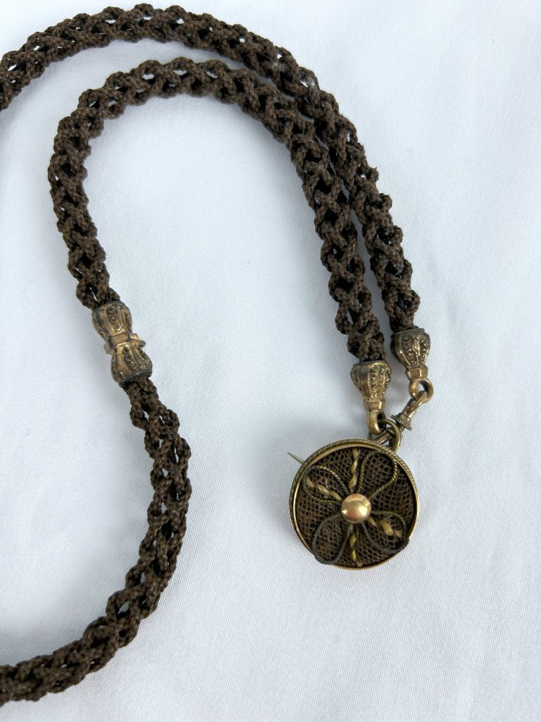 An image of a chain made from braided human hair with metal ornamentation and a round brooch with a floral design made from two different colors of human hair.