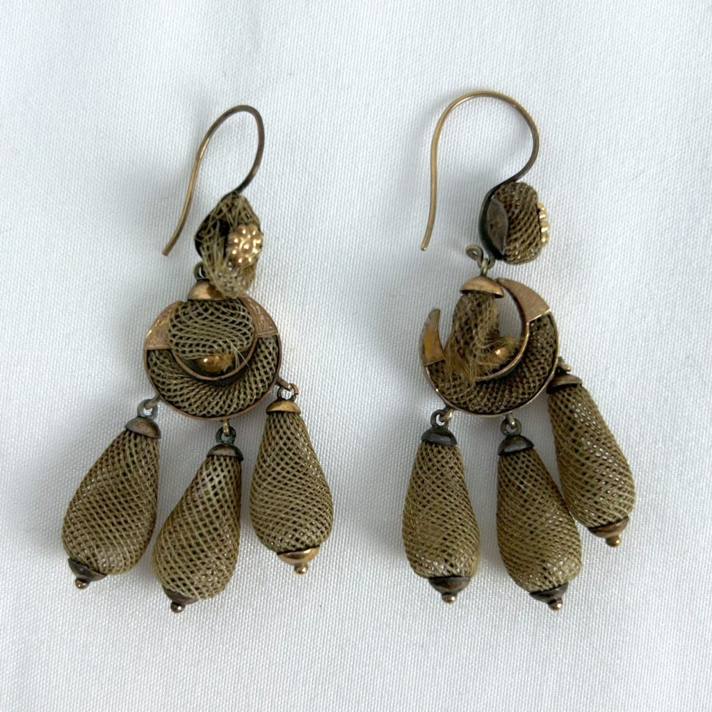 A photo of a pair of earrings made of metal and human hair that has been braided and shaped into 3 dimensional forms.