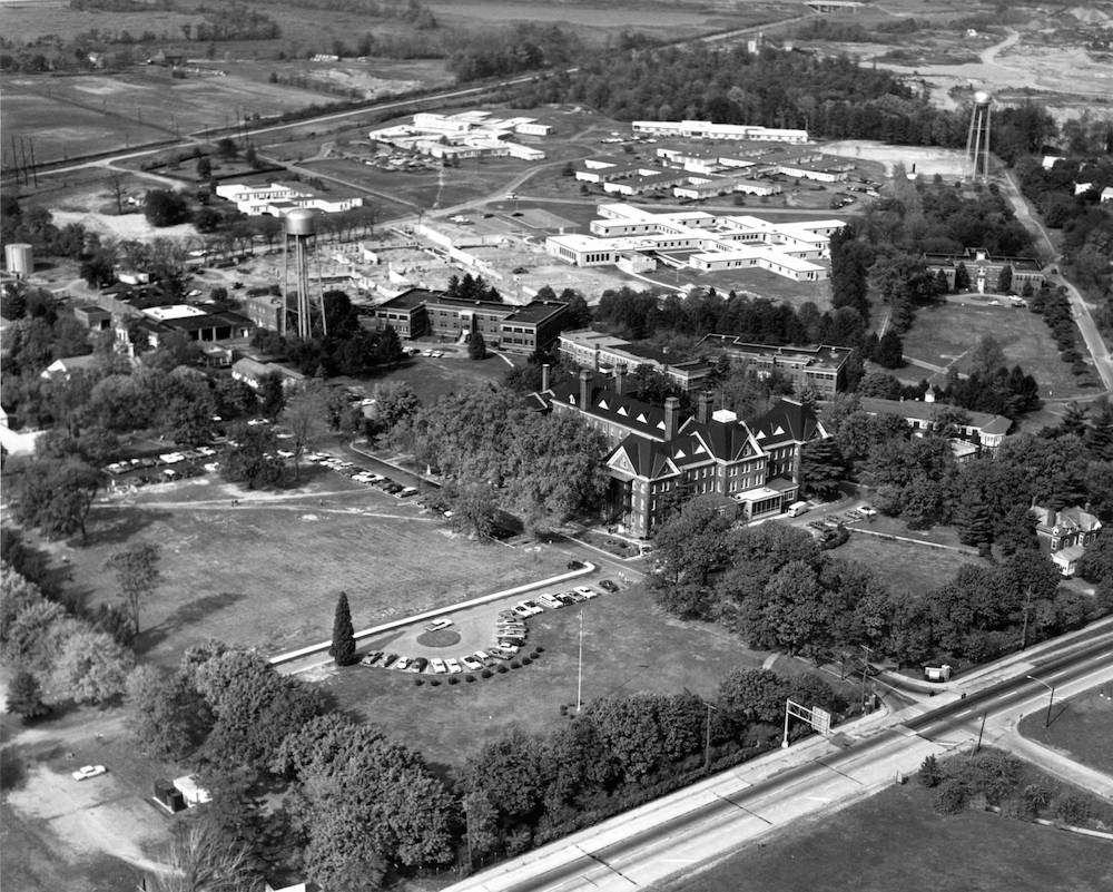 A black and white aerial photograph of a large building situated behind large trees.