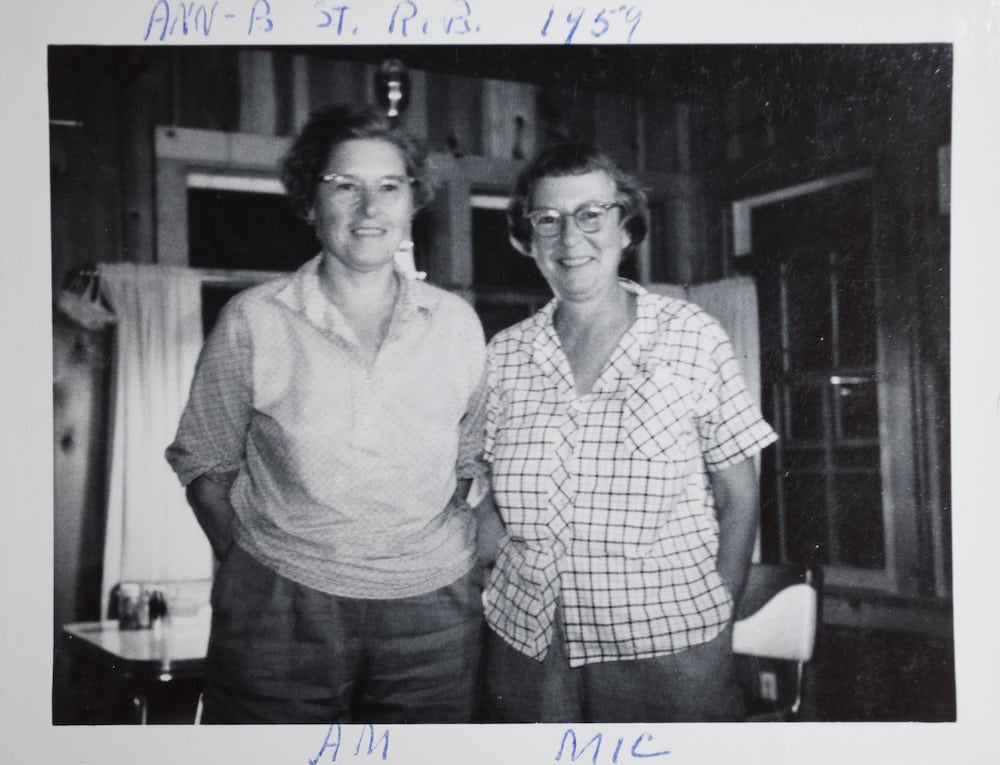 Two white women with short hair, glasses, and button-up shirts stand smiling in a kitchen. 