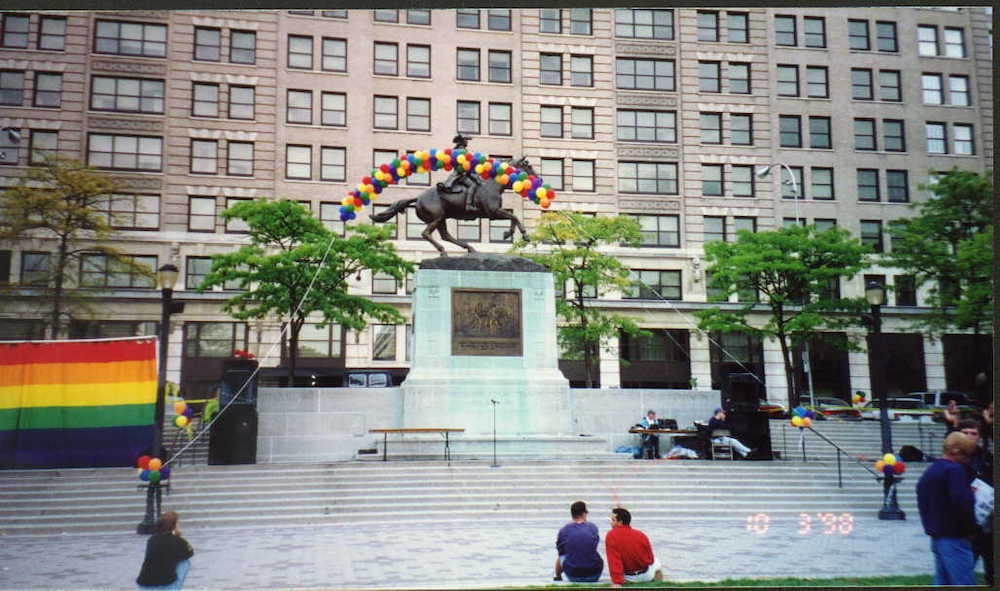 A rainbow flag and balloon arch is seen in a public square.