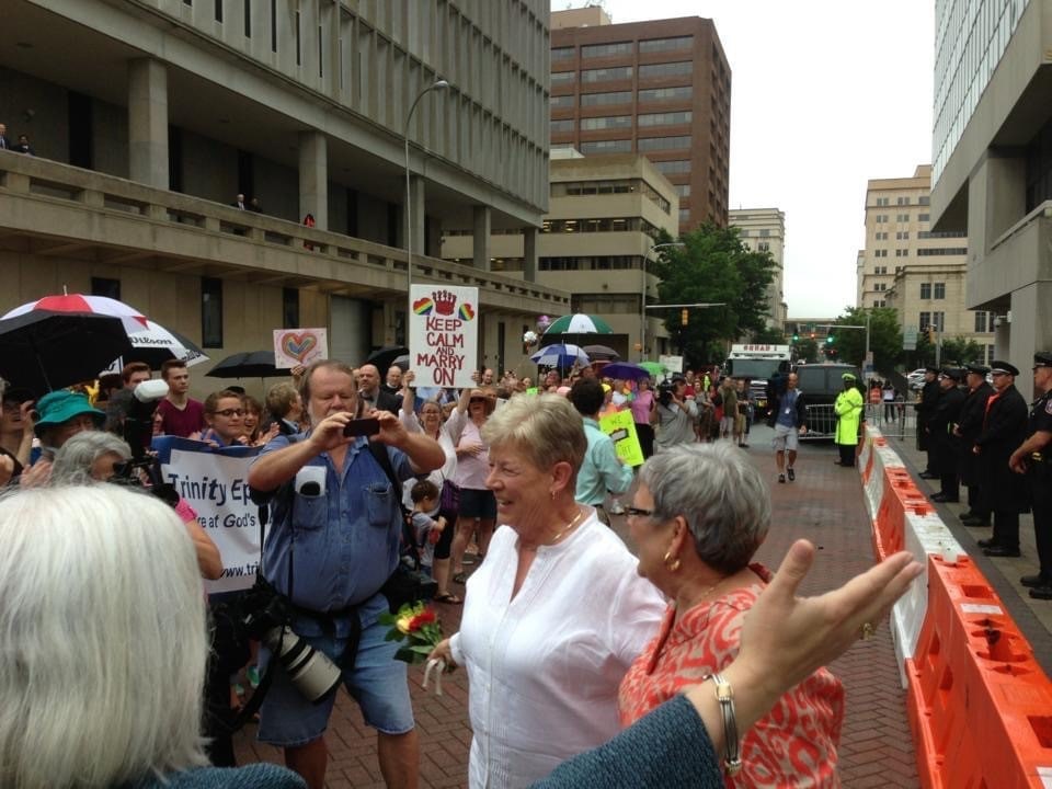 Two white women stand outside greeting a crowd of supporters. On the right are a line of police officers and barricades to protect the newlyweds.
