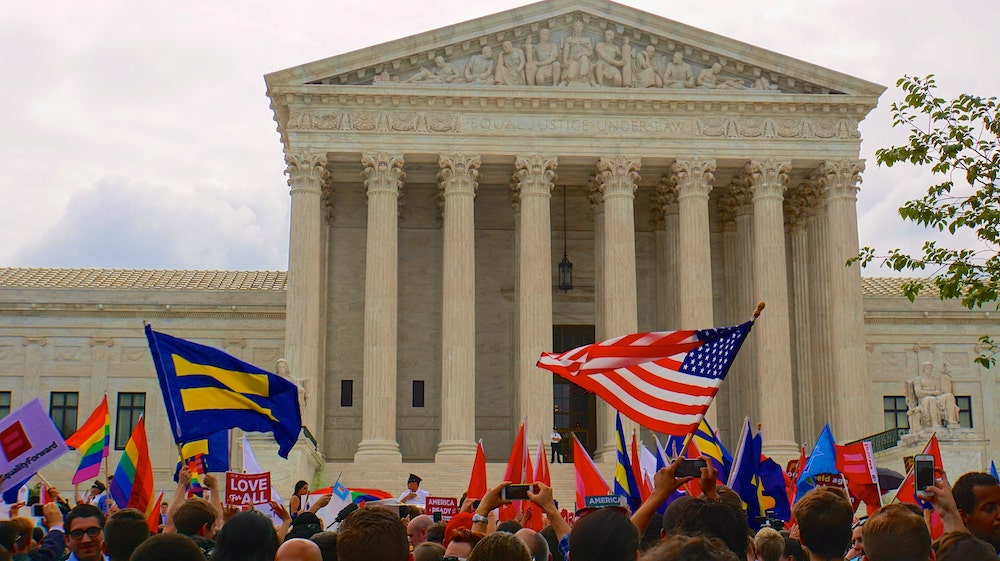 A crowd of people waving rainbow flags appear in front of the Supreme Court.