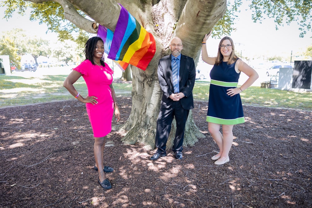 Three legislators, a black woman, a white man and a white woman, stand outside with a pride flag.