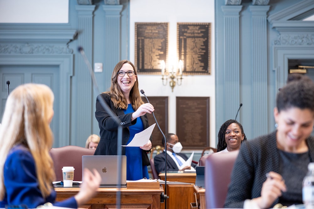 A white woman with long brown hair smiles as she stands on the senate floor. 