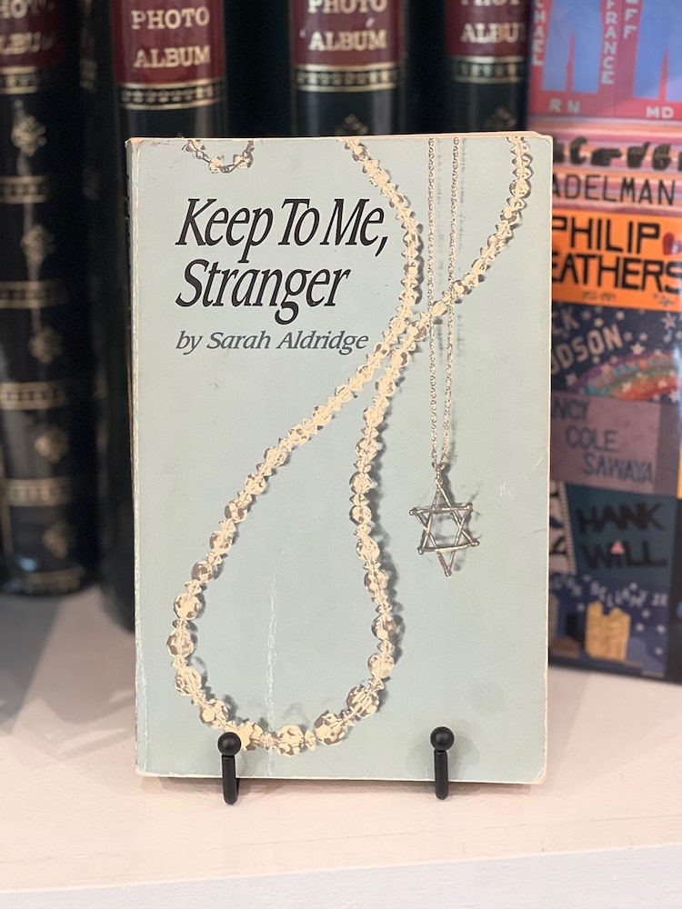 A paperback copy of the novel “Keep To Me, Stranger,” is displayed. The cover depicts two necklaces, a beaded necklace and the star of David