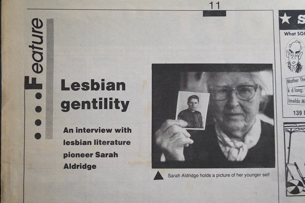 A photograph of an elderly white woman holding a photo of her younger self appears in a newspaper feature titled “Lesbian Gentility.”