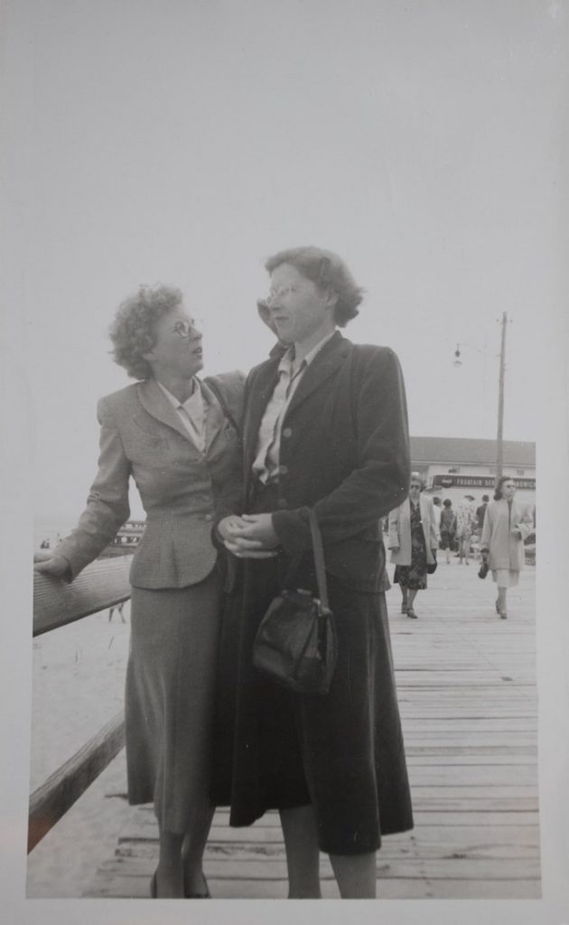 A black and white photograph of 2 white women on a boardwalk. Both women are wearing suit jackets, skirts and glasses.