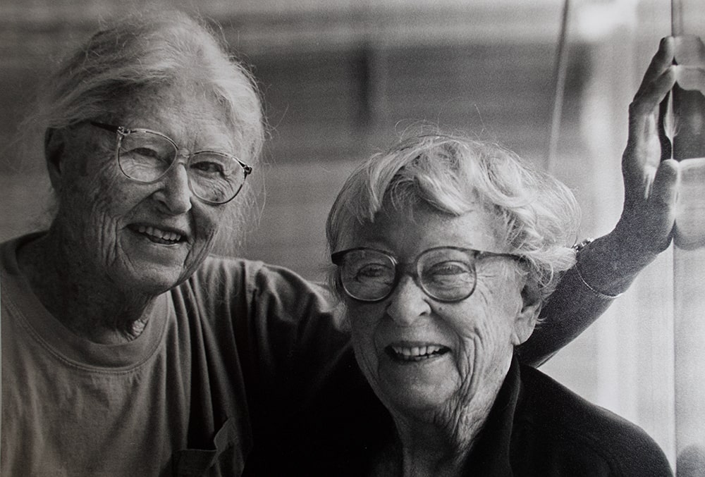 A black and white photograph of two elderly women wearing glasses and smiling.