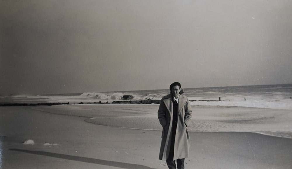 A black and white photograph of a white woman in a hat and long coat walking on the beach towards the viewer.