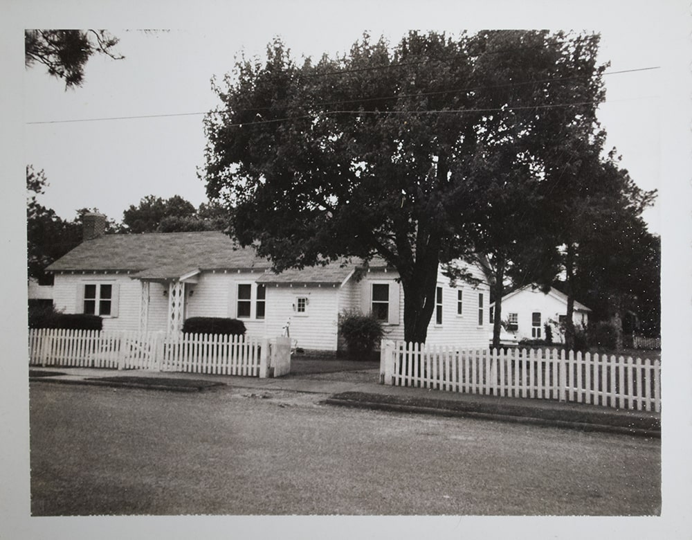 A black and white photograph of a white house behind a white france with a large tree in the front yard.