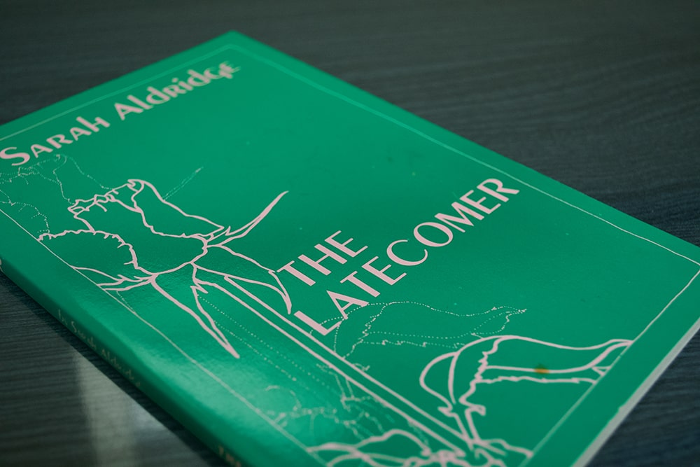 A green book with an illustration of a rose with the title “The Latecomer”