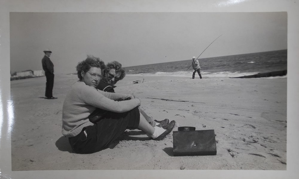 A black and white photograph of 2 white women sitting on the beach, facing the ocean, but are looking at the camera with straight faces.