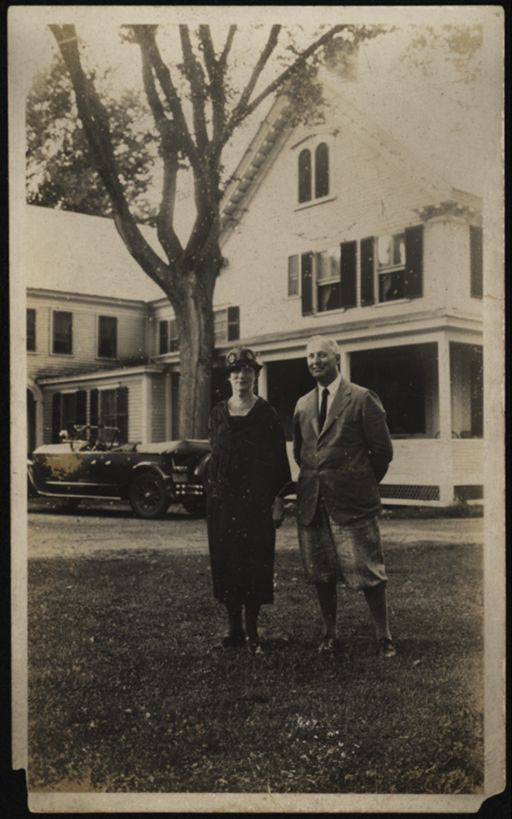 A white man and woman standing on a lawn in front of a white house, tree and car. 