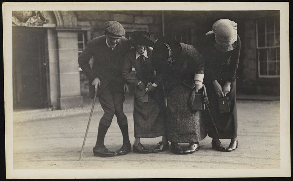 One man and three women playfully pose for a photograph outside. The man is holding a cane and his knees are turned in together.