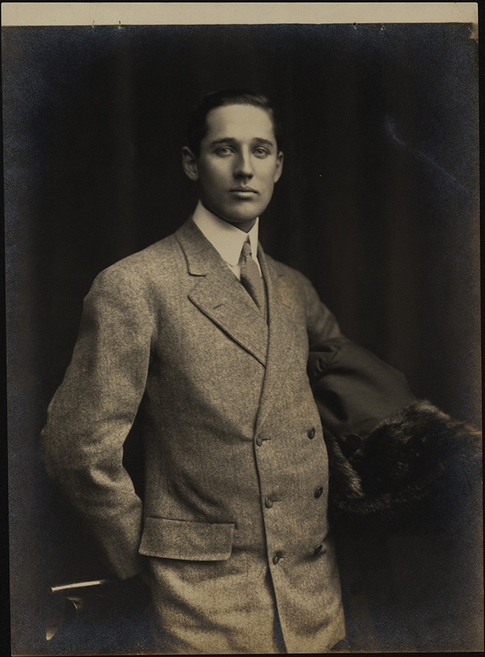 A white man with short brown hair looking straight at the camera. He wears a gray suit jacket with six buttons, a white shirt, and tie. 