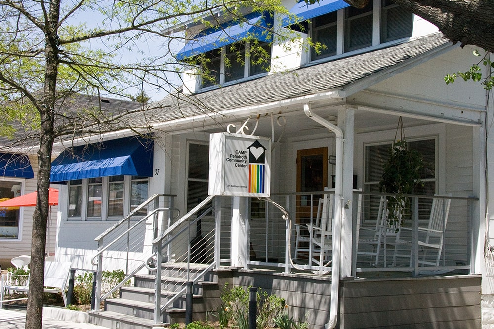 Five stairs lead to a covered porch with three white rocking chairs. A sign for CAMP Rehoboth Community Center hangs from the porch.