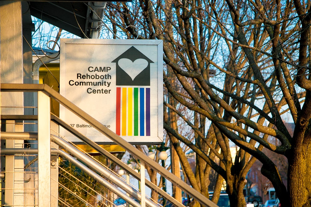 A square, white sign featuring a logo with a white heart inside of a black house with a vertical rainbow flag underneath.