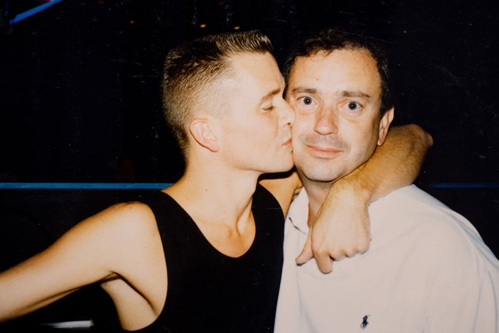 Two white men pose for a picture, one kissing the other’s cheek.