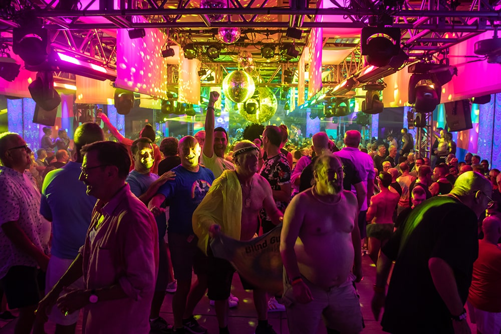 A crowd of men dancing in a large room with colorful lights and a disco ball.