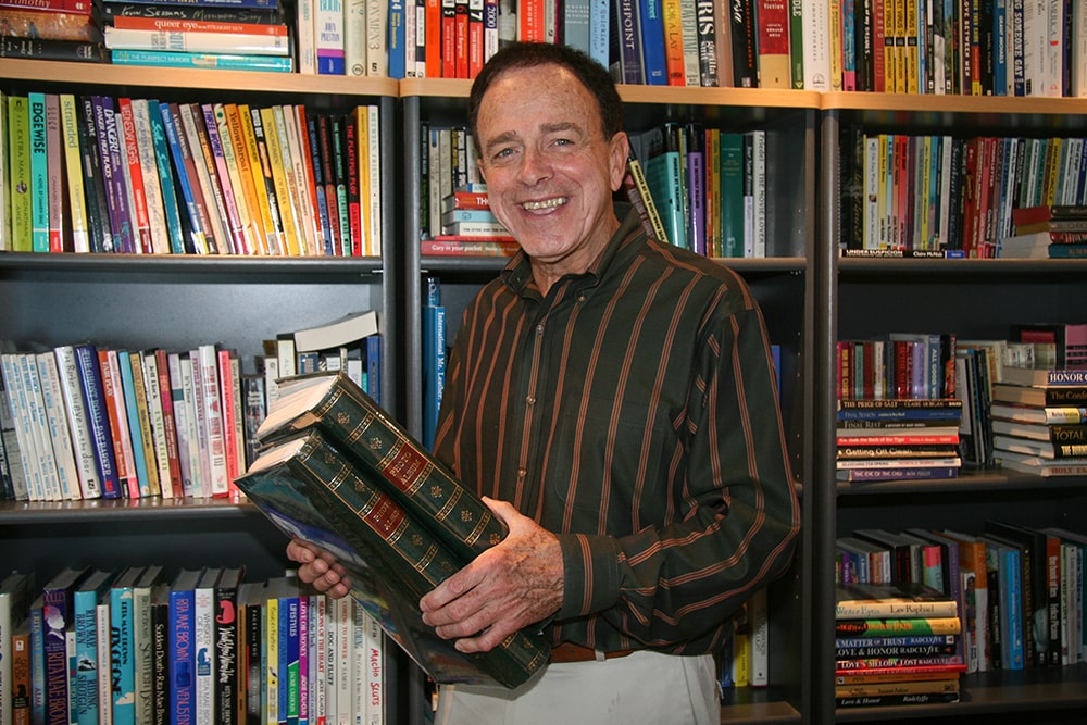 An elderly man with dark brown hair smiles for the camera, holding two large photo albums. He stands in front of three bookshelves filled with books.