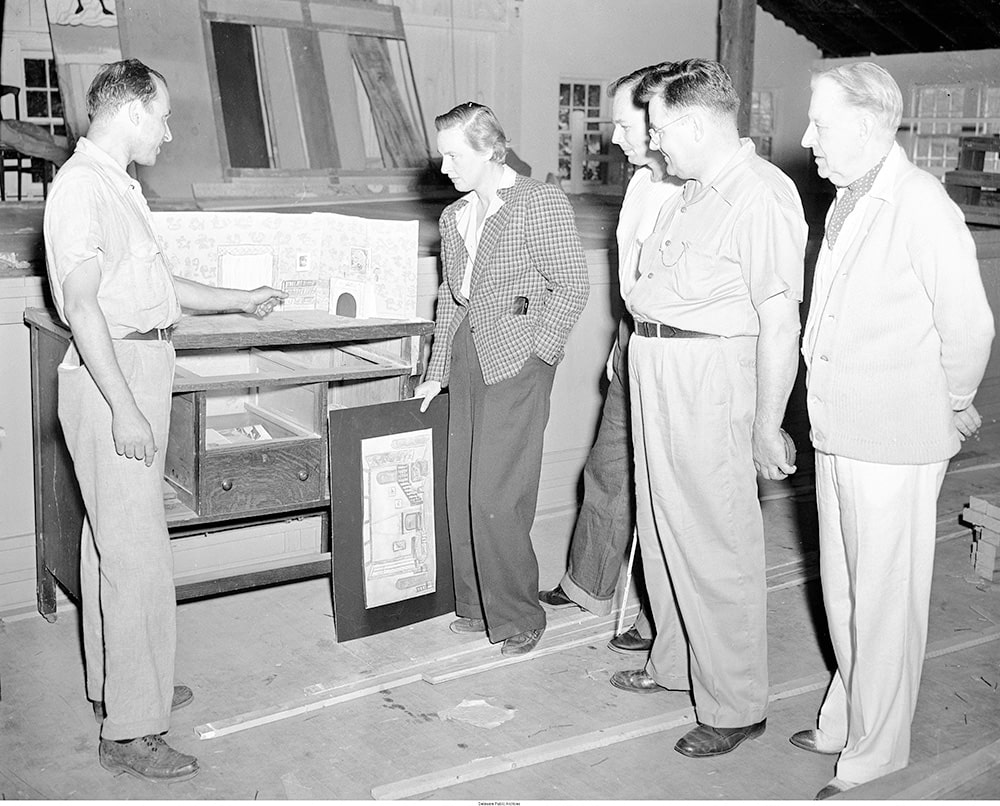 A woman and 4 men are seen looking at a model of a stage set. The woman is white, has her short hair styled in a masculine fashion, and she is wearing a plaid suit jacket and pants.