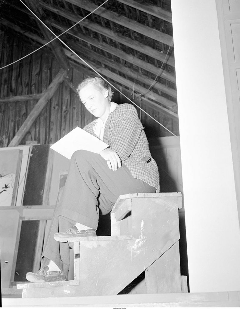 A woman in masculine attire is seen sitting down and reading a sheet of paper.