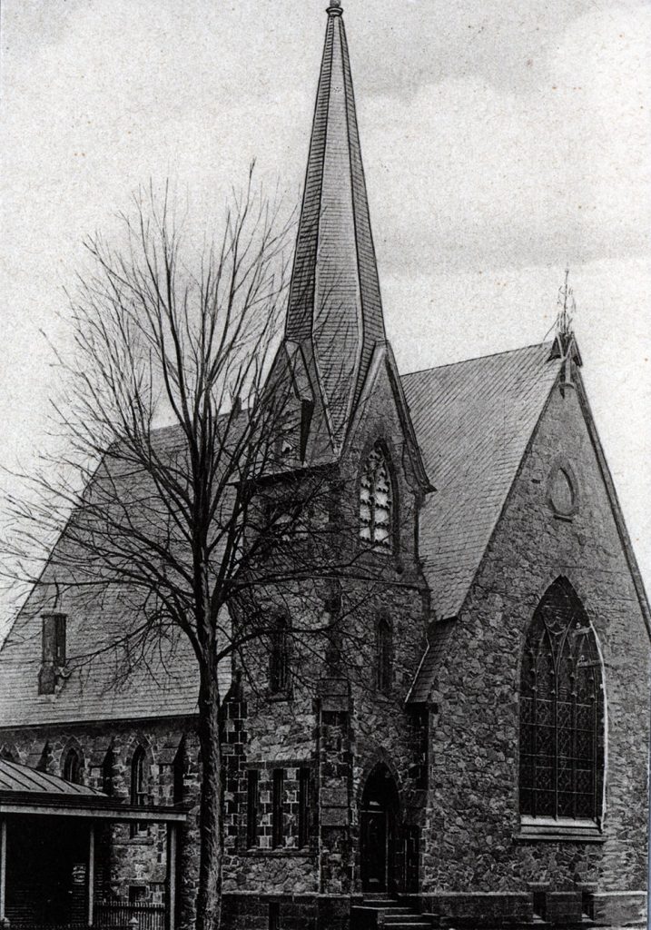 A black and white photograph of a church with a tall steeple, large pointed arches, and stained glass windows. 