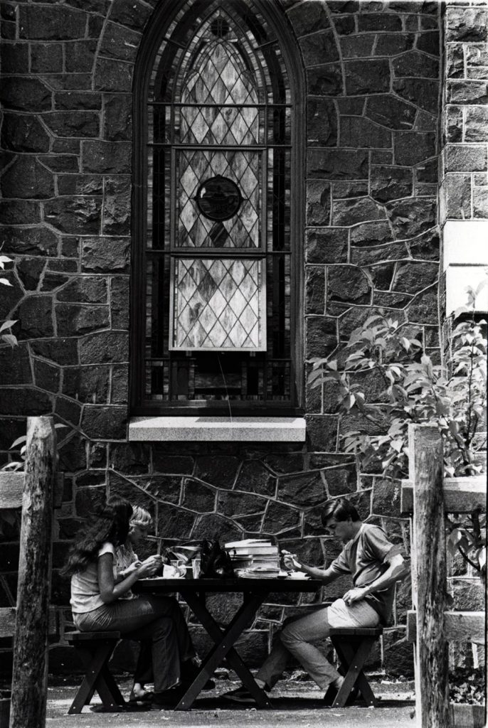 Two students, captured in profile, are seen sitting outside at a table under a stained glass window.