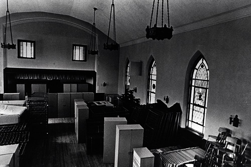 The interior of a hall with stained glass windows is seen filled with extra furniture, boxes and chairs.