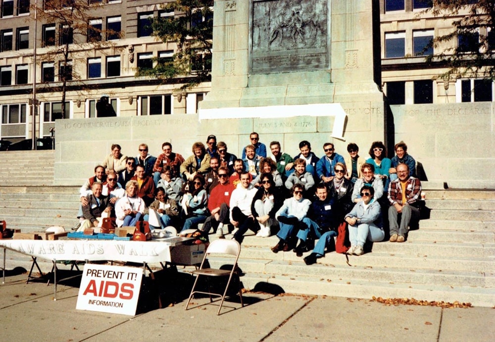 A group of activists sit on steps behind a table that reads “Delaware AIDS Walk.” A sign leaning on the table reads. “Prevent it! AIDS Information.”