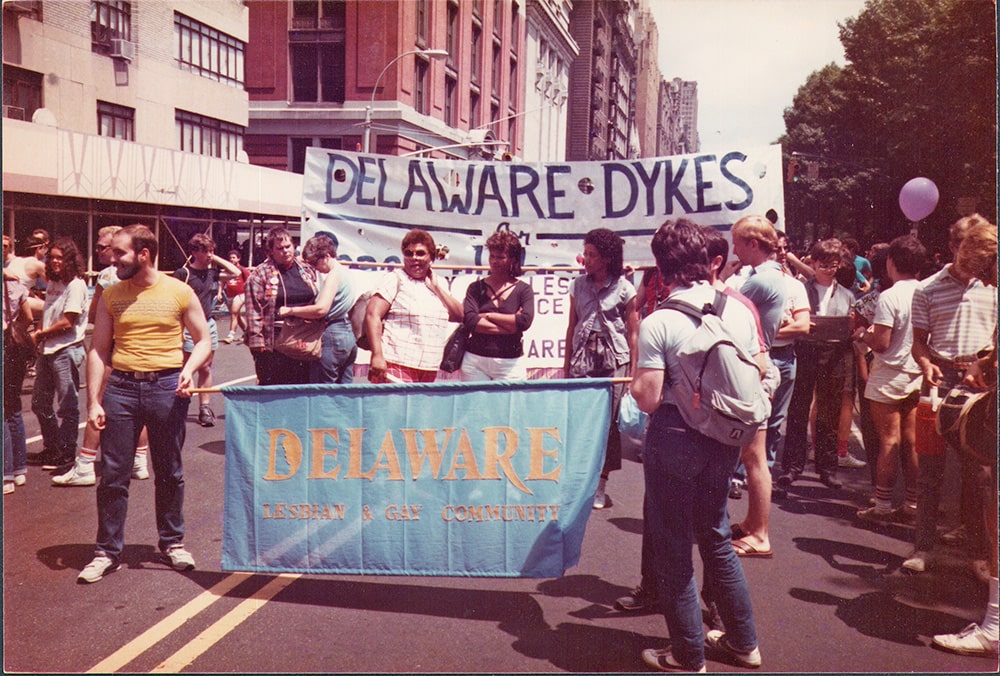 People stand in the street holding two signs. One reads, “Delaware Lesbian & Gay Community.” The other partially reads, “Delaware Dykes.”