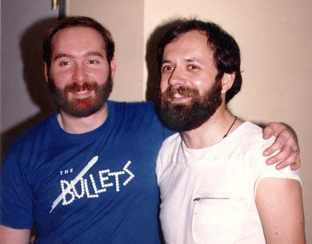 Two light-skinned men with beards and mustaches smile with their arms around each other.