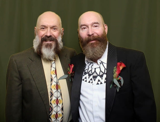 Two light-skinned men with full beards and mustaches smile for the camera. Both men are wearing suit jackets with red flowers pinned to the lapels.