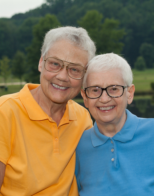 Two white women with short white hair and glasses smile for a photograph in a park.