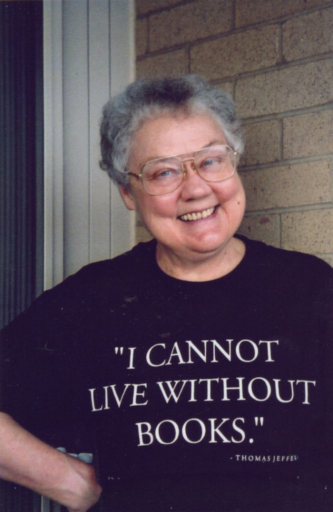 A white woman with short gray hair and glasses, smiles at the camera while wearing a shirt that says “I Cannot Live without Books- Thomas Jefferson.”