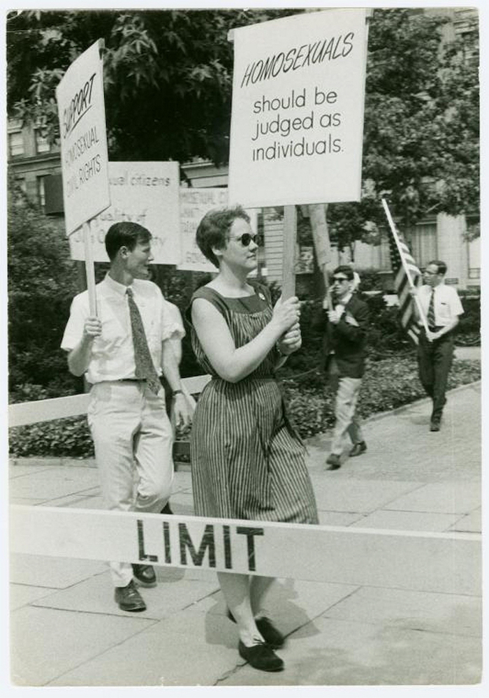 A white woman with short hair wearing a dress, marches in a picket line, holding a sign that reads, “Homosexuals should be judged as individuals.”