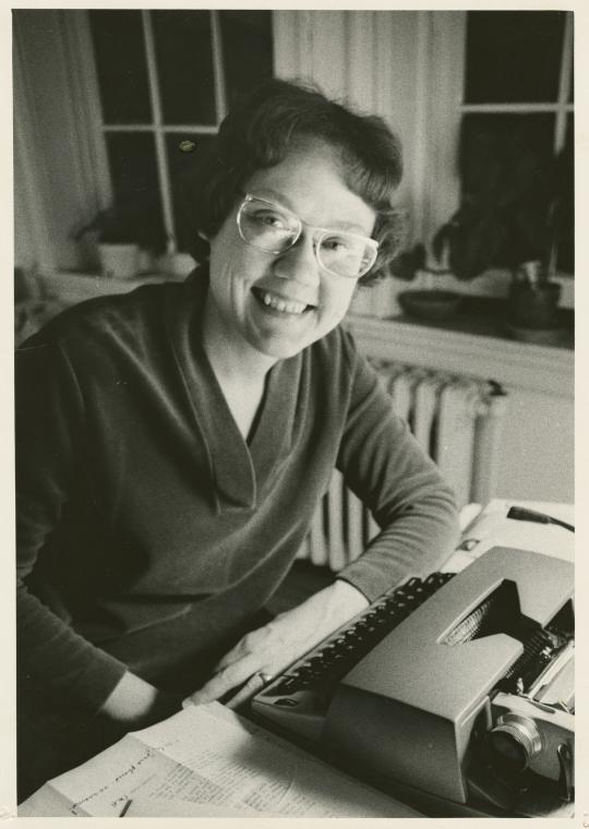 A white woman with short brown hair sits at a table in front of a typewriter, smiling at the camera.