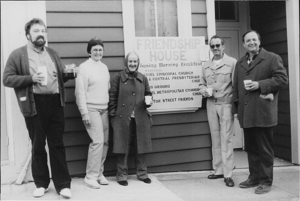 A group of five people stand outside next to a sign reading “Friendship House, Sunday Morning Breakfast.”