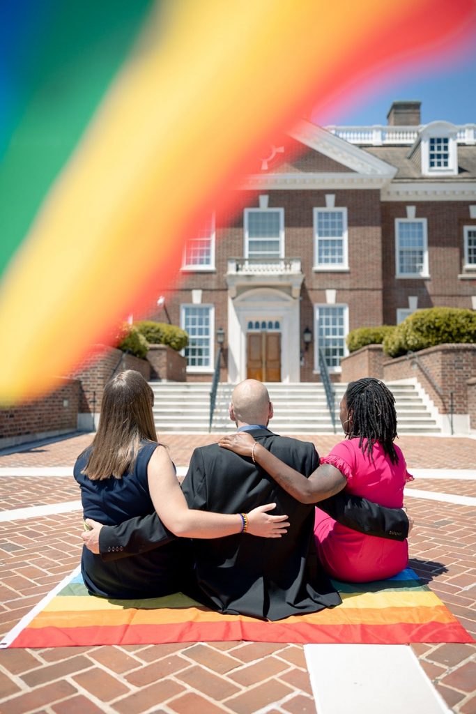 Three legislators are seen from behind, sitting on a rainbow flag, looking toward the legislative hall.