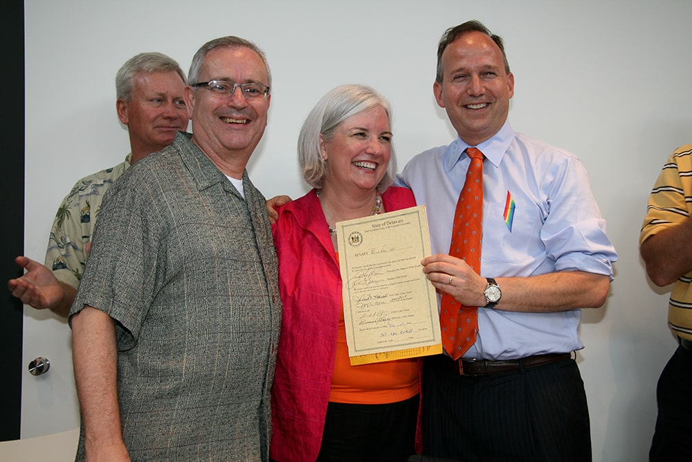 Three white people smile for the camera with a State of Delaware bill.