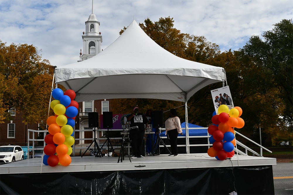 Two Black women are seen on a stage addressing an audience. The woman with a microphone wears a shirt that says “Black Trans Lives Matter.”