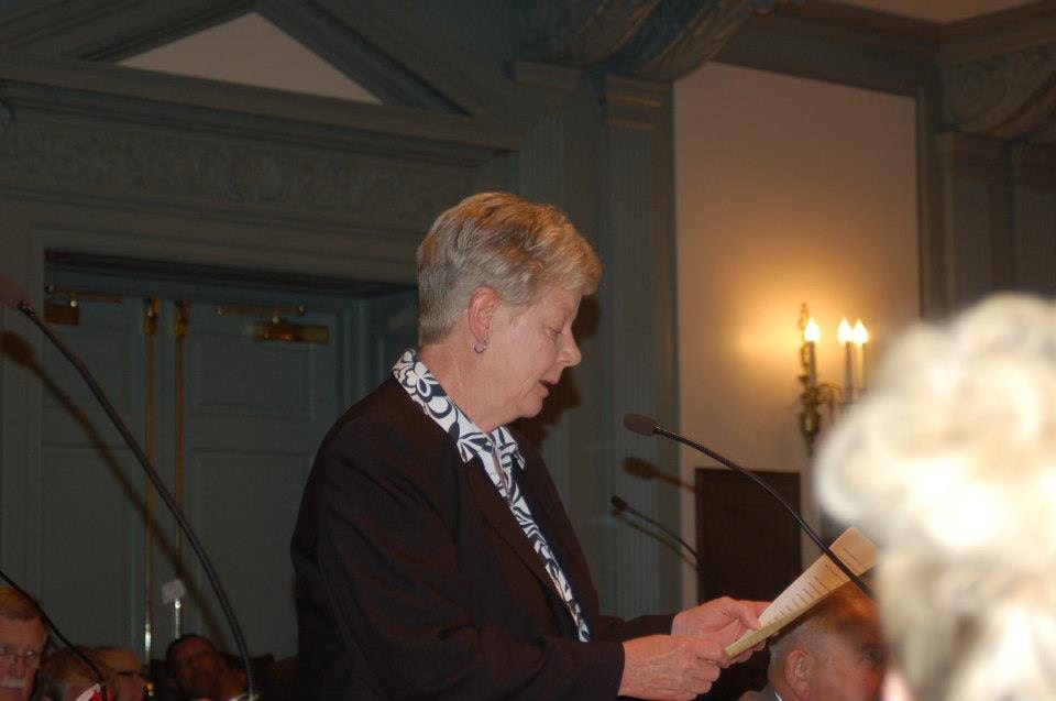 A white woman with short blonde hair is seen reading her testimony with a microphone on the House floor.