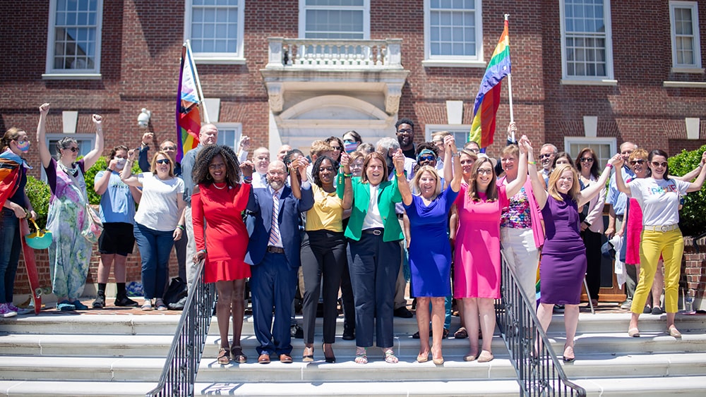 A group of legislators wearing colorful outfits stand together, holding hands, in front of Legislative Hall.