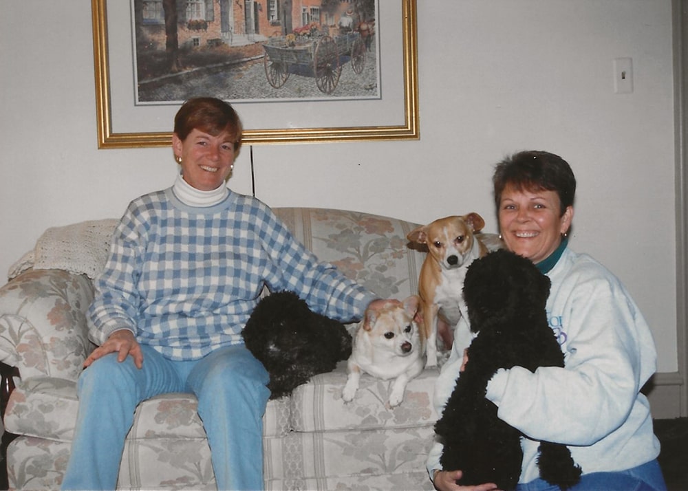 Two white women with short hair pose with 4 dogs on a floral couch
