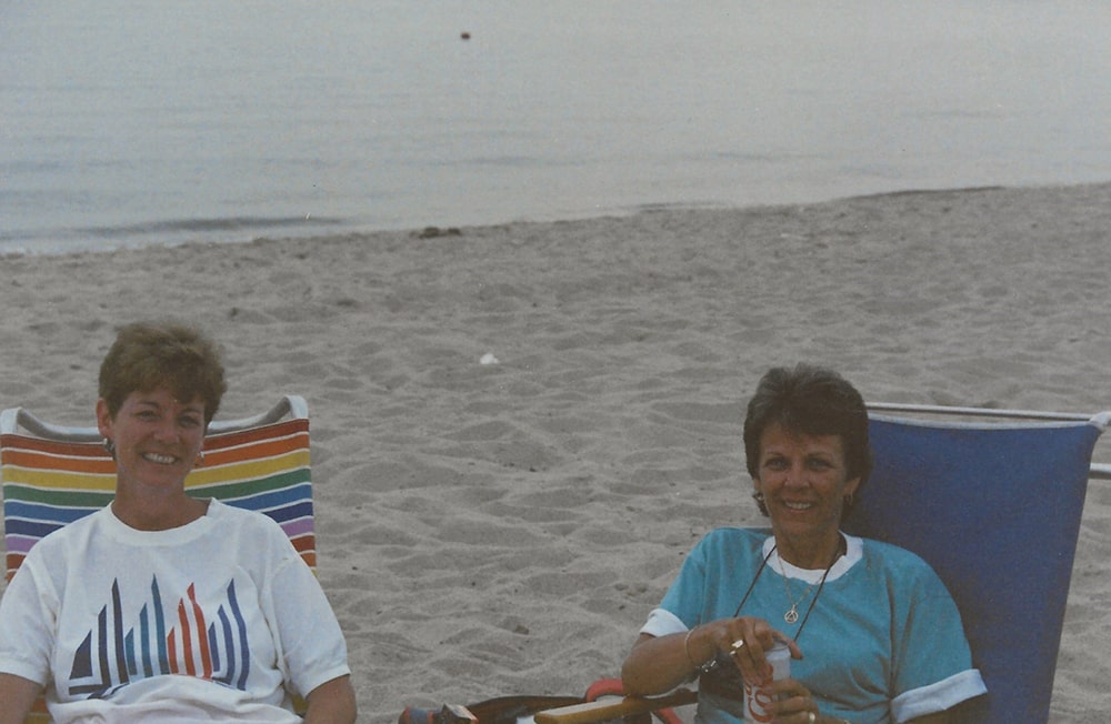 Two white women with short hair are seated on the beach near the ocean in beach chairs.