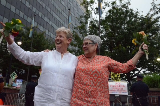 Two white women each carry bouquets of flowers as they greet supporters outside. Protesters, police and barricades can be seen in the background.